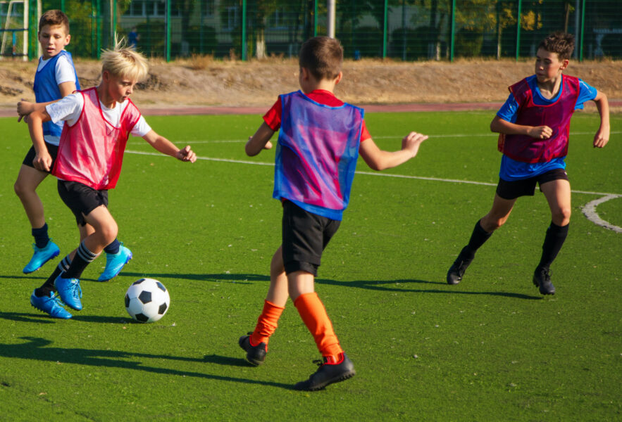 The photo shows a group of young soccer players in colorful jerseys on a green turf field. They are actively playing and competing for the ball, with some players in motion and others watching.